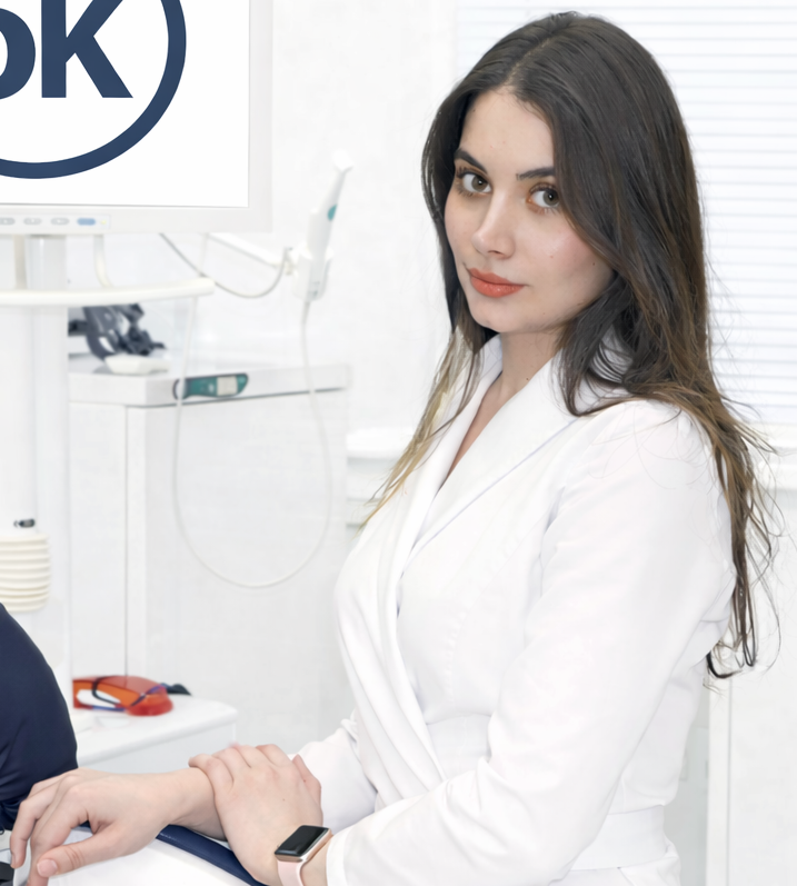 Доктор клиники ОртоКапа — врач-стоматолог, хирург-имплантолог Зарета Замбековна Хасуева в белом халате на фоне стоматологической клиники